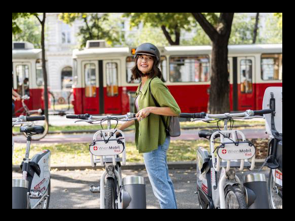 junge Frau steht mit Fahrradhelm bei der WienMobil Rad Station. Im Hintergrund fährt eine Straßenbahn vorbei.