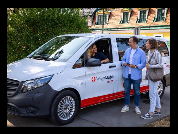 eine Frau sitzt in einem Kastenwagen mit der Aufschrift Wien Mobil Hüpfer sie lächelt zwei Personen zu die neben dem Fahrzeug stehen